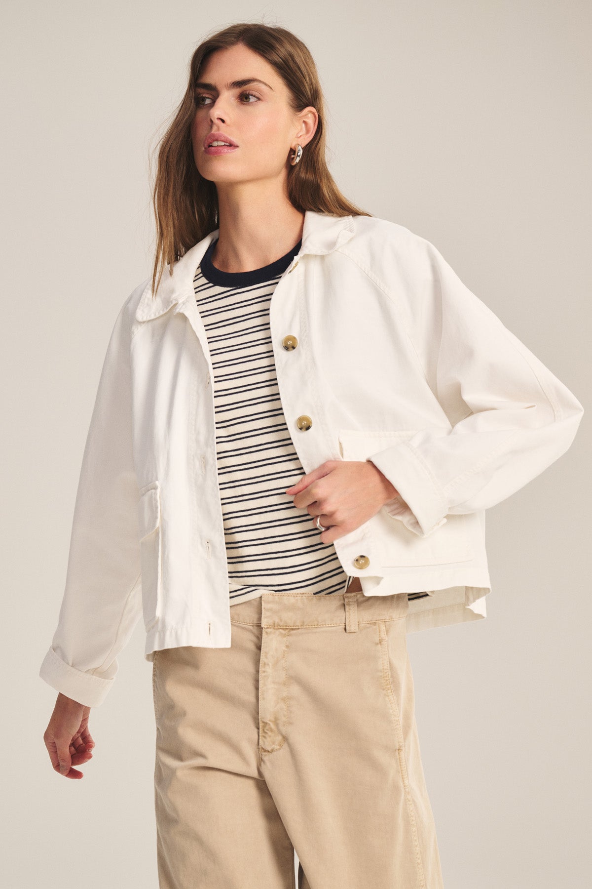 A woman wears the CARRINGTON JACKET by Velvet by Graham & Spencer over a black-and-white striped shirt and beige pants, standing against a plain background.
