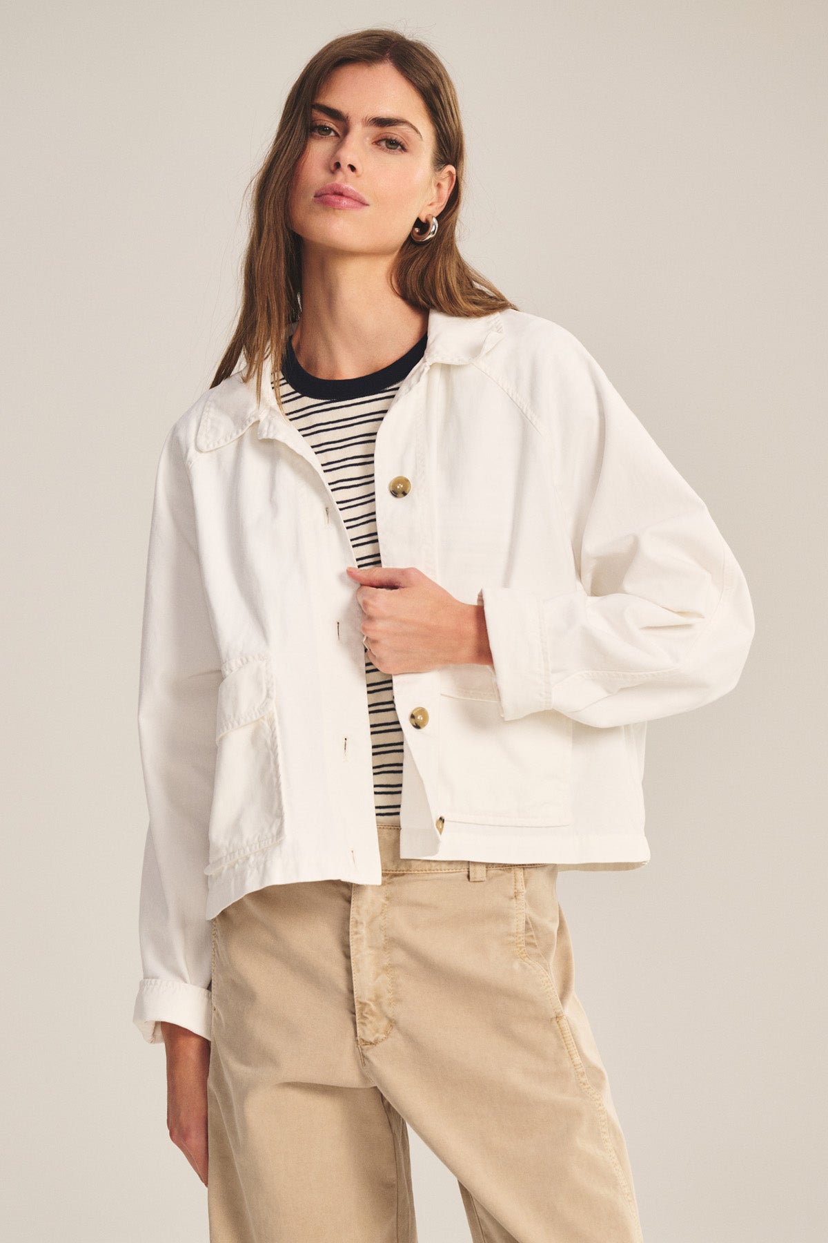 A woman wears the CARRINGTON JACKET by Velvet by Graham & Spencer over a black-and-white striped shirt and beige pants, standing against a neutral background.