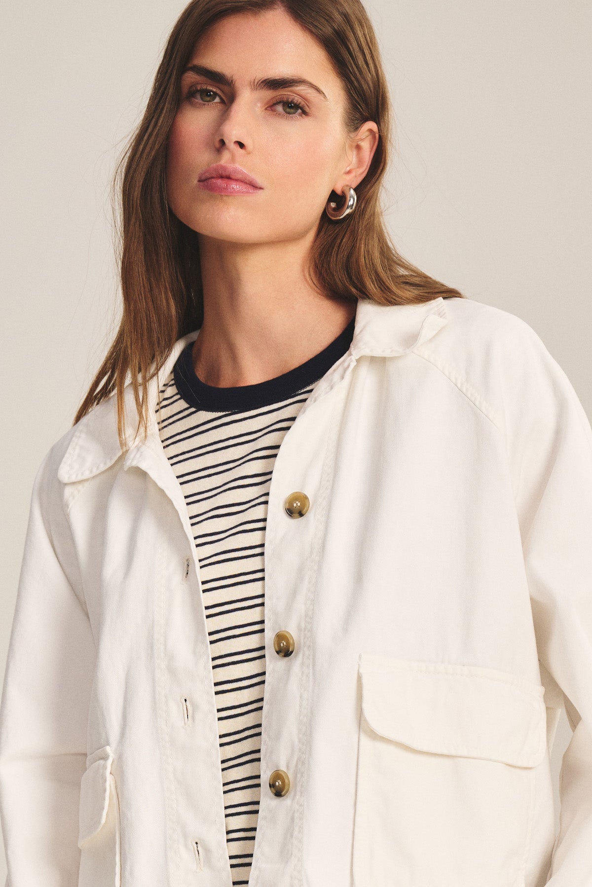 A woman with long brown hair models the CARRINGTON JACKET by Velvet by Graham & Spencer—a white cotton twill layer worn over a black-and-white striped shirt, perfect for versatile styling against a neutral backdrop.-40909714587841