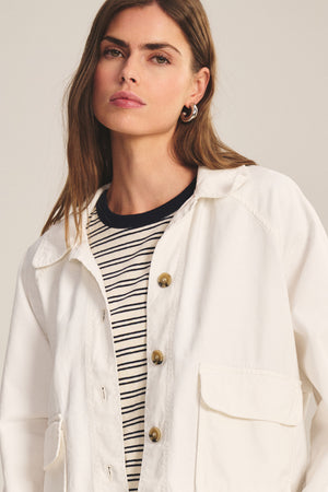 A woman with long brown hair models the CARRINGTON JACKET by Velvet by Graham & Spencer—a white cotton twill layer worn over a black-and-white striped shirt, perfect for versatile styling against a neutral backdrop.