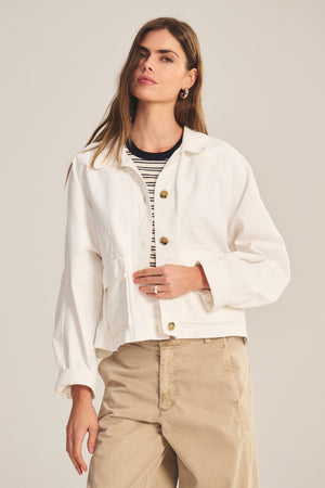 A woman stands against a plain background, wearing the CARRINGTON JACKET by Velvet by Graham & Spencer over a striped shirt and beige pants.