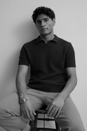 A man with short curly hair sits on a stool against a plain wall, wearing the Velvet by Graham & Spencer CARSTON SWEATER POLO, light pants, and a wristwatch.