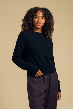 A person with curly hair stands against a plain beige background, looking slightly to the side, wearing Velvet by Graham & Spencer’s ALBA CASHMERE SWEATER—a soft knit black cashmere piece featuring a classic crew neckline—paired with dark pants.