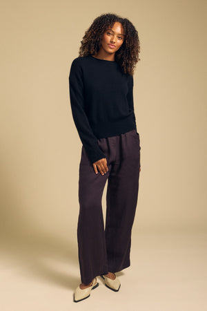 A woman stands against a beige background wearing the ALBA CASHMERE SWEATER by Velvet by Graham & Spencer, featuring a classic crew neckline, paired with loose dark brown pants and cream pointed shoes, with her hands in her pockets.