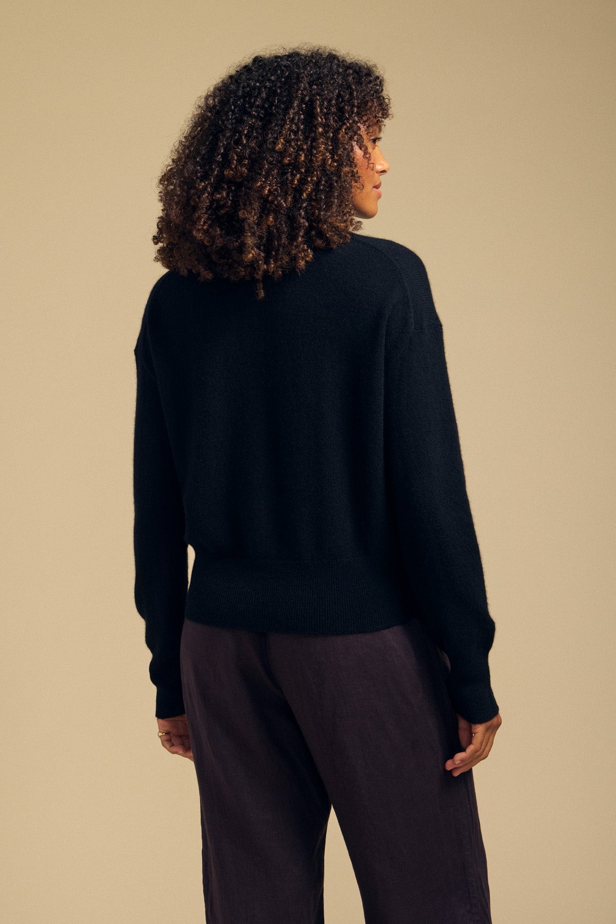 A person with curly hair is seen from behind against a plain beige background, wearing the ALBA CASHMERE SWEATER by Velvet by Graham & Spencer— a black, soft knit crewneck sweater—paired with dark pants.