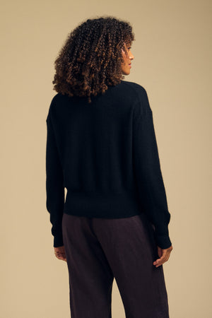A person with curly hair is seen from behind against a plain beige background, wearing the ALBA CASHMERE SWEATER by Velvet by Graham & Spencer— a black, soft knit crewneck sweater—paired with dark pants.