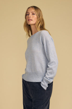 A person stands with hands in pockets, wearing the ALBA CASHMERE SWEATER by Velvet by Graham & Spencer—a light blue sweater featuring a classic crew neckline—paired with dark pants and posed against a plain beige background.