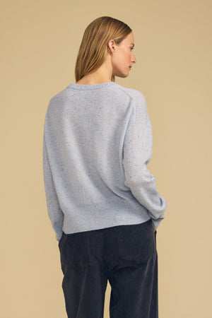 A woman with straight, light brown hair is shown from the back, wearing the ALBA CASHMERE SWEATER by Velvet by Graham & Spencer in light blue, featuring a classic crew neckline and paired with dark pants against a beige background—an effortless look that captures everyday indulgence.