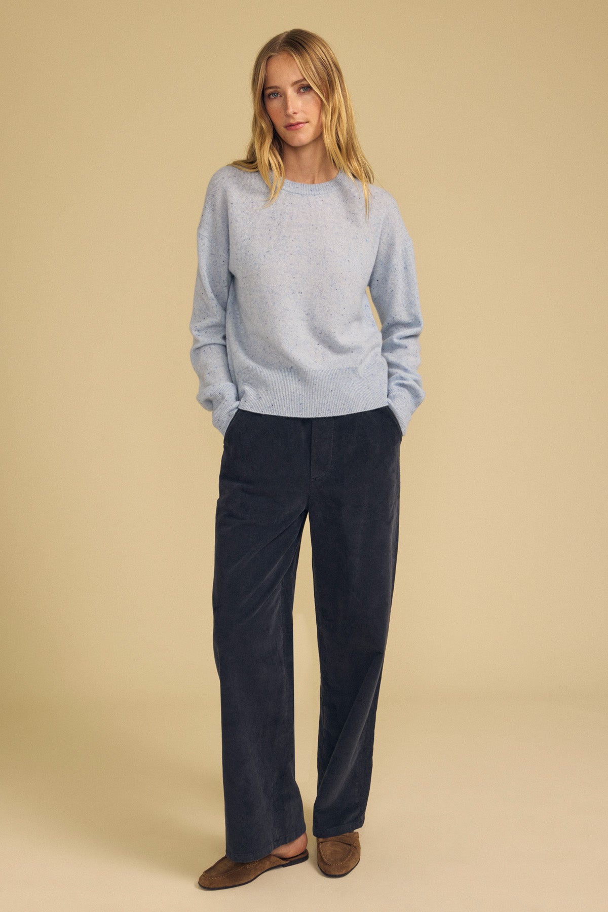 A person stands against a beige background wearing the ALBA CASHMERE SWEATER by Velvet by Graham & Spencer—a light blue cashmere sweater with a classic crew neckline—paired with dark blue wide-leg pants and brown shoes, hands in pockets.
