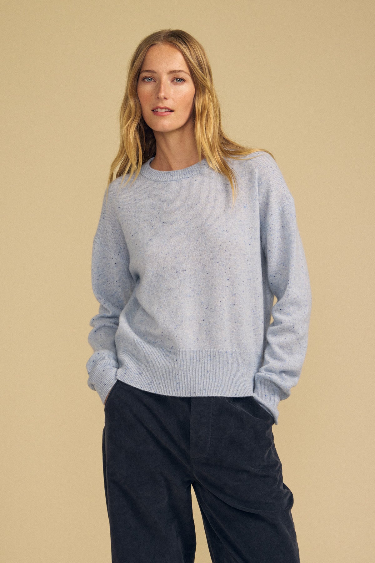 A person with long blonde hair wears the ALBA CASHMERE SWEATER by Velvet by Graham & Spencer—a light blue speckled cashmere crewneck—paired with dark pants, standing against a plain beige background with hands in their pockets.
