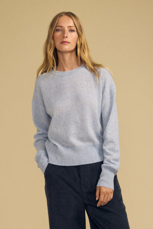 A person stands against a beige background, wearing Velvet by Graham & Spencer’s ALBA Cashmere Sweater in light blue—a classic crewneck paired with dark pants. Their relaxed posture and neutral expression capture the understated luxury of this everyday indulgence.