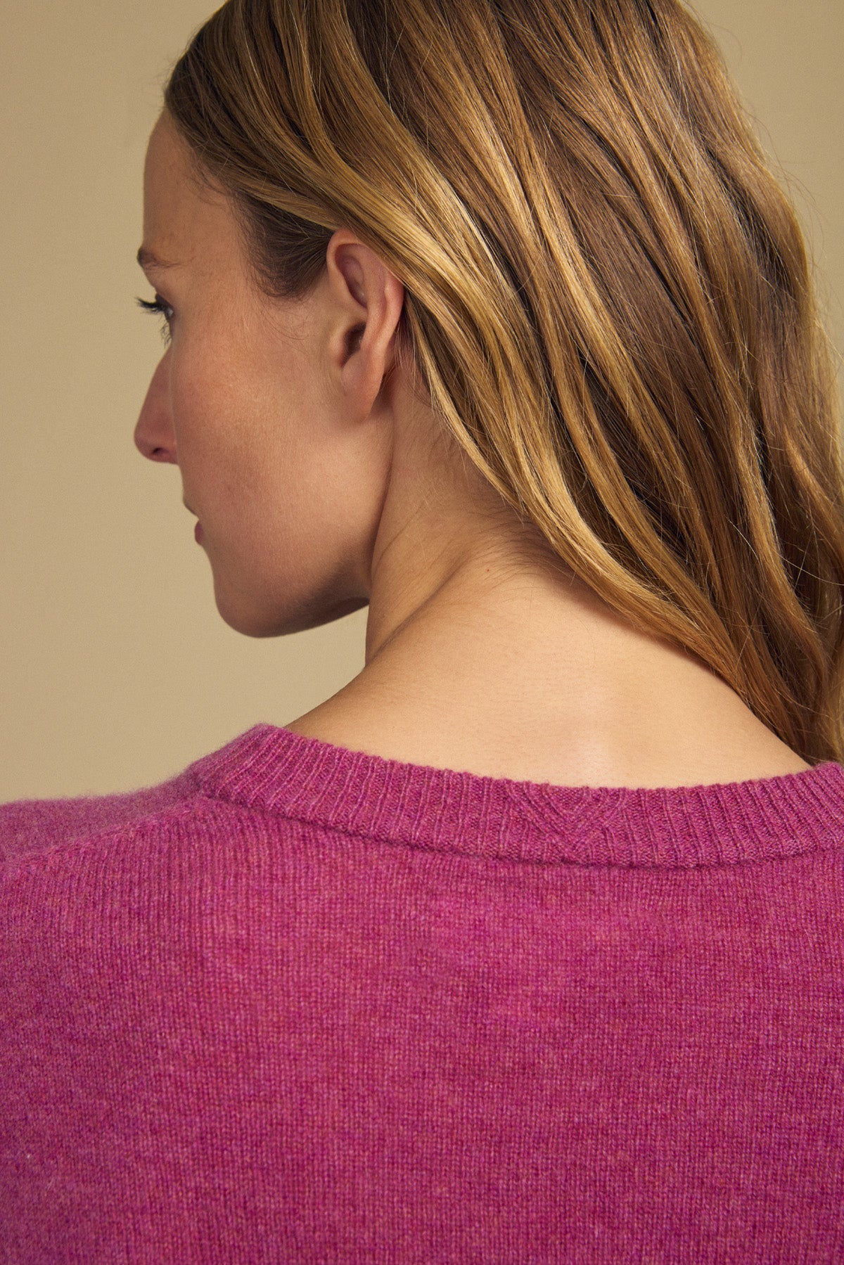 A woman with light brown hair is shown from behind, wearing the ALBA CASHMERE SWEATER by Velvet by Graham & Spencer—a purple cashmere sweater featuring a classic crew neckline—against a neutral background.
