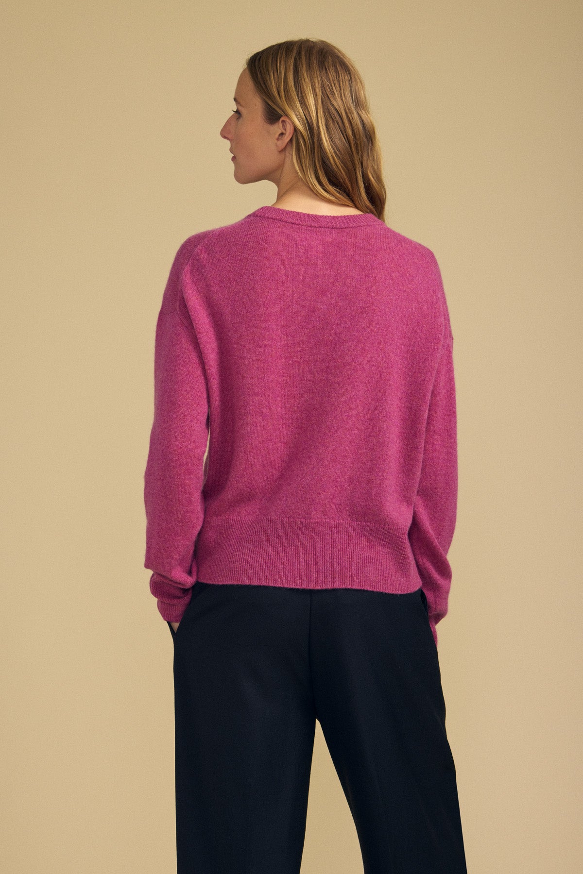 A person stands with their back to the camera, wearing the ALBA CASHMERE SWEATER by Velvet by Graham & Spencer—a pink 100% cashmere crewneck—paired with black pants against a beige background.-42063714582721
