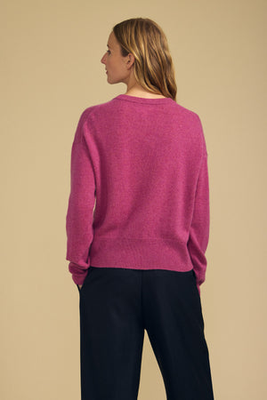 A person stands with their back to the camera, wearing the ALBA CASHMERE SWEATER by Velvet by Graham & Spencer—a pink 100% cashmere crewneck—paired with black pants against a beige background.