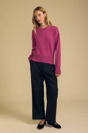 A person stands against a beige background wearing the Velvet by Graham & Spencer ALBA CASHMERE SWEATER in pink, featuring a crew neckline, paired with wide-leg black pants and black flats, with one hand in their pocket.
