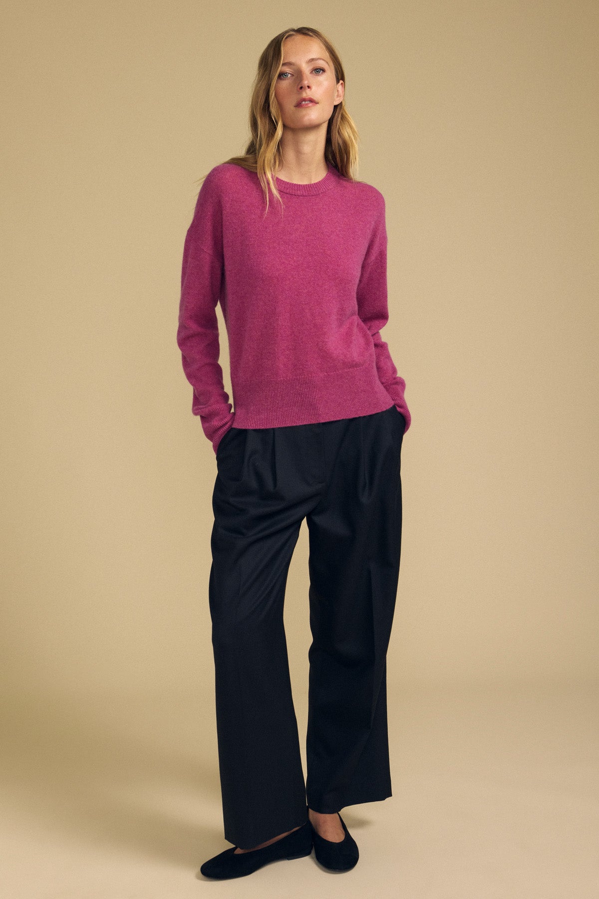A person stands against a beige background wearing the Velvet by Graham & Spencer ALBA Cashmere Sweater, featuring a pink hue and crew neckline, paired with loose black pants and black shoes, hands in pockets.