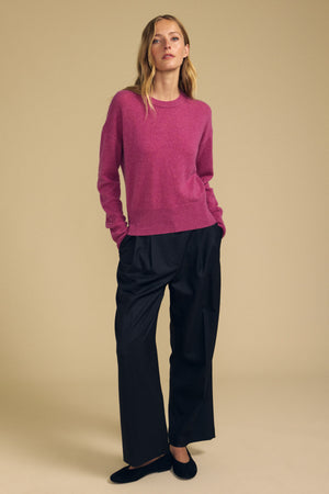 A person stands against a beige background wearing the Velvet by Graham & Spencer ALBA Cashmere Sweater, featuring a pink hue and crew neckline, paired with loose black pants and black shoes, hands in pockets.
