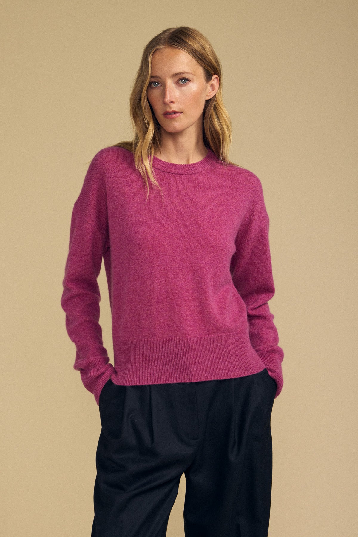 A person wears the ALBA CASHMERE SWEATER by Velvet by Graham & Spencer—a long-sleeve pink cashmere style with a classic crew neckline—paired with loose black pants, standing against a plain beige background and gazing slightly to the side with hands in their pockets.
