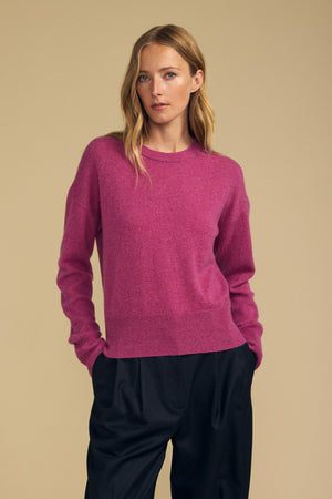 A person wears the ALBA CASHMERE SWEATER by Velvet by Graham & Spencer—a long-sleeve pink cashmere style with a classic crew neckline—paired with loose black pants, standing against a plain beige background and gazing slightly to the side with hands in their pockets.