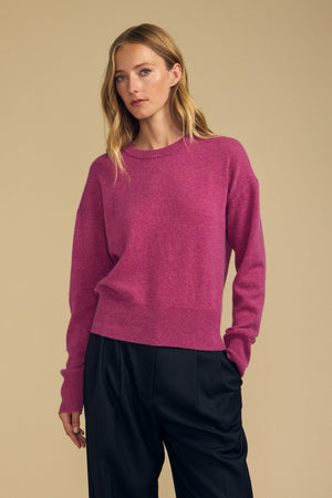 A person stands against a beige background wearing the ALBA CASHMERE SWEATER by Velvet by Graham & Spencer—a pink 100% cashmere piece featuring a classic crew neckline—paired with loose black pants and one hand casually in their pocket.