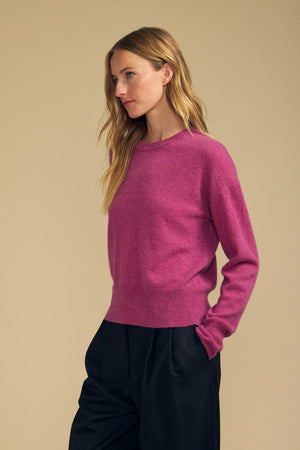 A woman with long, wavy blonde hair wears the ALBA CASHMERE SWEATER by Velvet by Graham & Spencer—a pink 100% cashmere sweater featuring a classic crew neckline—paired with black pants, as she stands against a plain beige background.