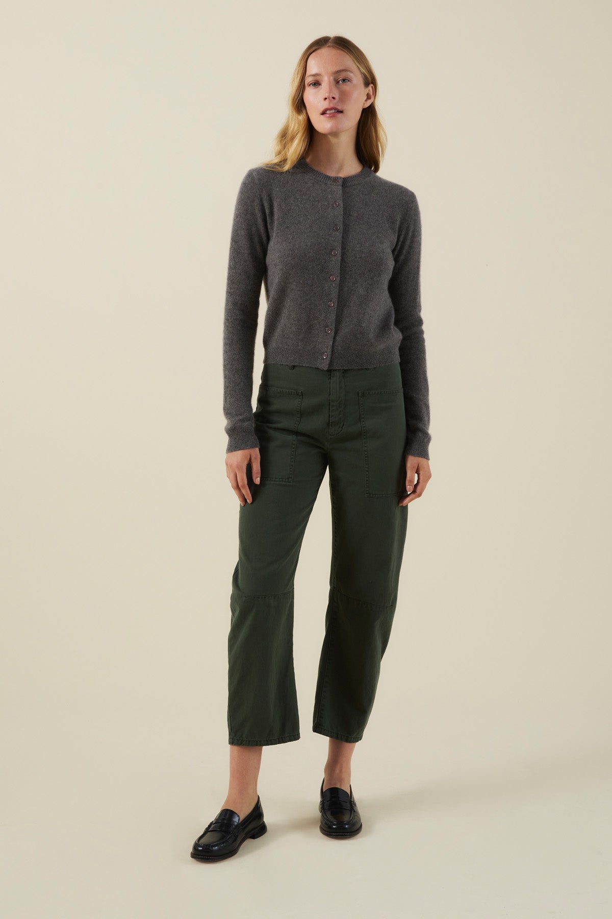 A person wearing a grey cropped AYLINA CASHMERE CARDIGAN, dark green utility pants, and black loafers.