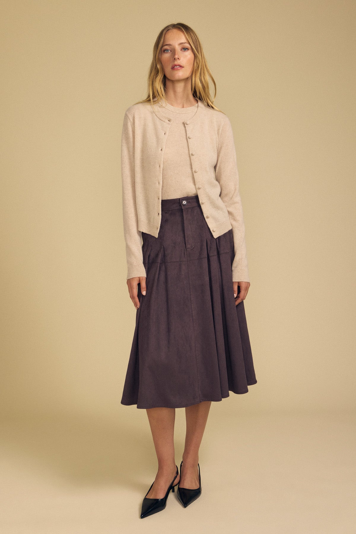 A woman stands against a beige background wearing a light tan cardigan with a coordinating top, styled with the BELINDA FAUX SUEDE SKIRT by Velvet by Graham & Spencer in dark brown—an ideal look for transitional style—finished with black pointed shoes.-41935336210625