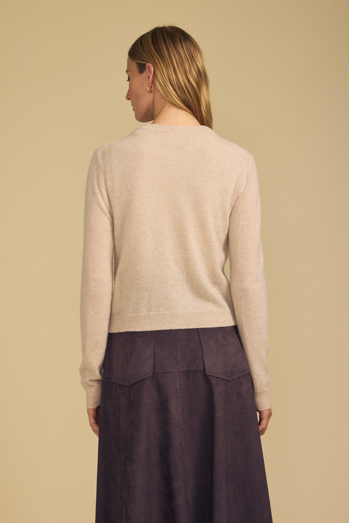 A person stands facing away, wearing the AYLINA CASHMERE CARDIGAN by Velvet by Graham & Spencer—a light beige women’s layering sweater—paired with a high-waisted dark purple skirt, and their straight light brown hair draped over one shoulder against a plain background.