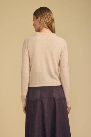 A person stands facing away, wearing the AYLINA CASHMERE CARDIGAN by Velvet by Graham & Spencer—a light beige women’s layering sweater—paired with a high-waisted dark purple skirt, and their straight light brown hair draped over one shoulder against a plain background.