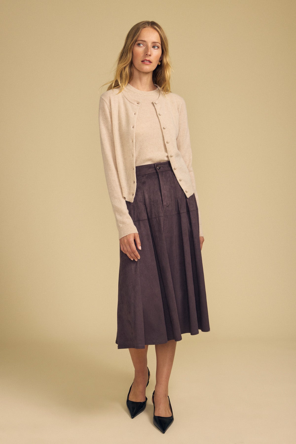 A woman stands against a beige background, effortlessly layering the AYLINA CASHMERE CARDIGAN by Velvet by Graham & Spencer—a light beige, cropped cashmere cardigan—over a matching top, paired with a brown midi skirt and black pointed-toe shoes.
