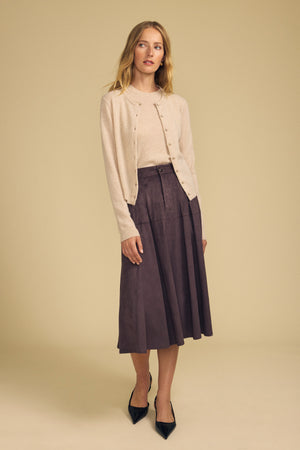 A woman stands against a beige background, effortlessly layering the AYLINA CASHMERE CARDIGAN by Velvet by Graham & Spencer—a light beige, cropped cashmere cardigan—over a matching top, paired with a brown midi skirt and black pointed-toe shoes.