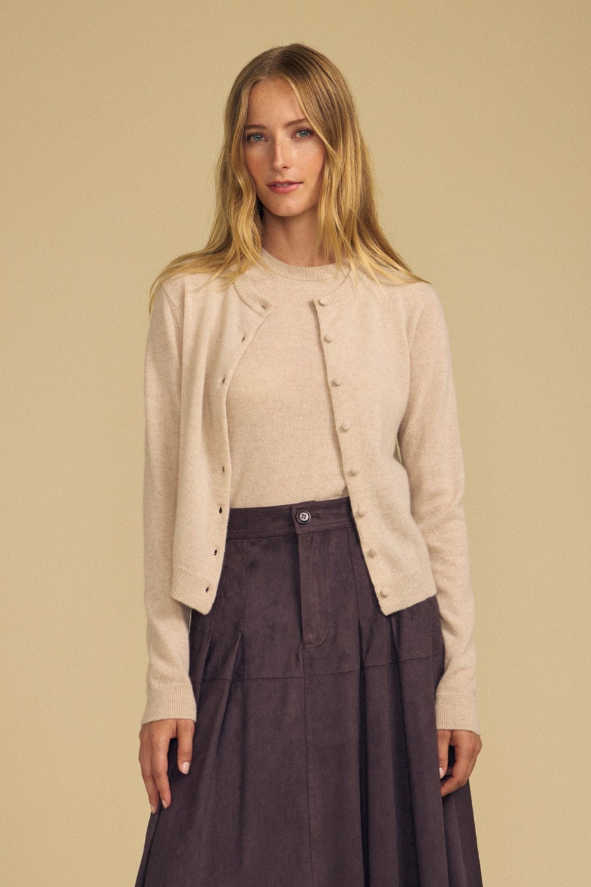 A woman stands against a beige background wearing the AYLINA CASHMERE CARDIGAN by Velvet by Graham & Spencer over a matching sweater, embracing stylish layering with a dark brown skirt.-41935336308929
