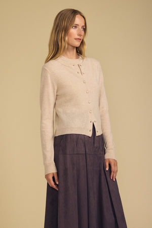 A woman wearing the AYLINA CASHMERE CARDIGAN by Velvet by Graham & Spencer—a beige, buttoned cardigan—paired with a long dark brown skirt, stands against a plain beige background.