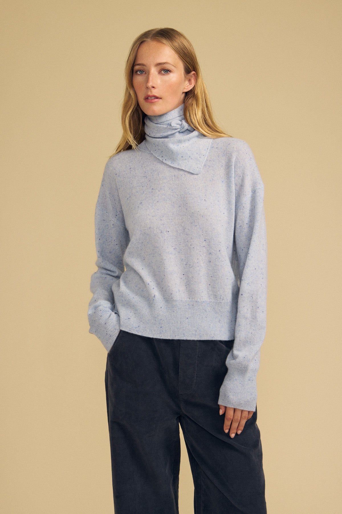 A woman stands against a plain beige background, wearing a light blue turtleneck sweater with long sleeves and dark trousers, styled with the HELEN CASHMERE SCARF by Velvet by Graham & Spencer as a chic triangular layering accessory.-42063740108993