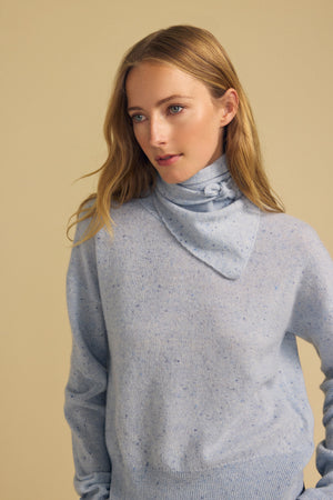 A woman with long blonde hair wears the Velvet by Graham & Spencer HELEN Cashmere Scarf—a light blue speckled triangular scarf—paired with a matching sweater, standing against a beige background.