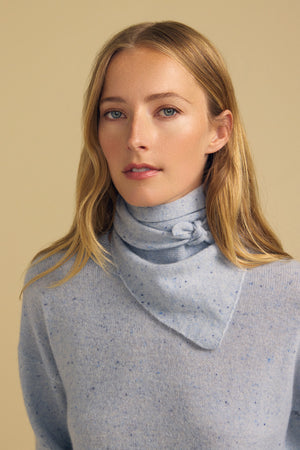 A woman with long blonde hair wears the HELEN CASHMERE SCARF by Velvet by Graham & Spencer, paired with a light blue speckled sweater, standing against a beige background.