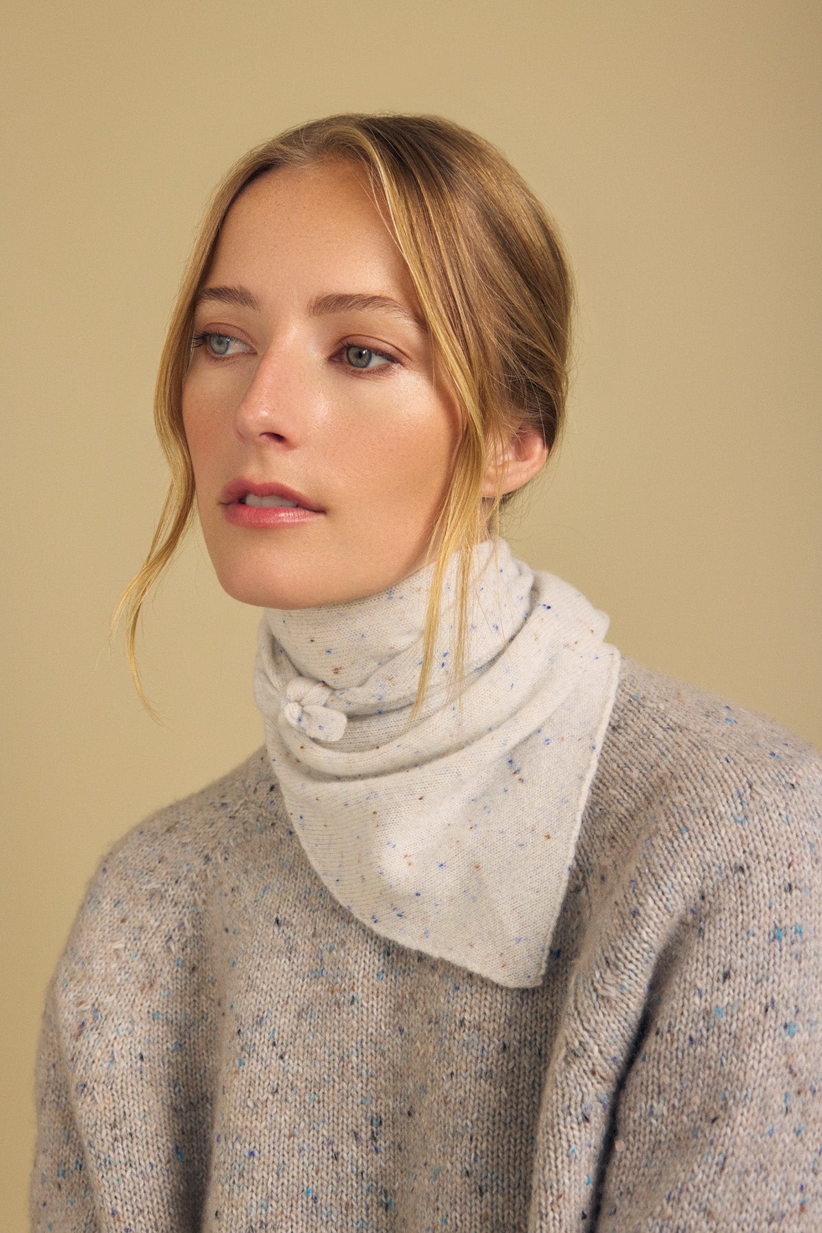 A woman with blonde hair in a low bun models the HELEN CASHMERE SCARF by Velvet by Graham & Spencer, styled as a chic buttoned neck warmer that matches her beige speckled sweater. She looks slightly to the side against a plain beige background.