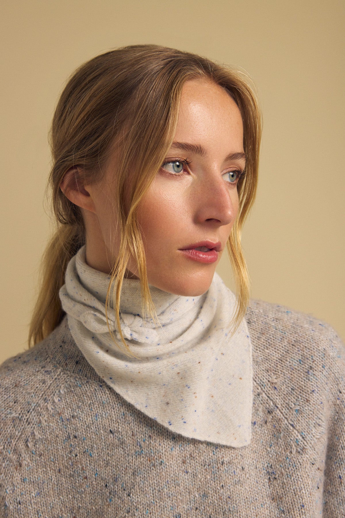 A woman with blonde hair in a low ponytail wears the HELEN CASHMERE SCARF by Velvet by Graham & Spencer—a light, speckled triangular accessory—layered over a beige speckled sweater, as she looks slightly to the side against a plain beige background.-41960847311041