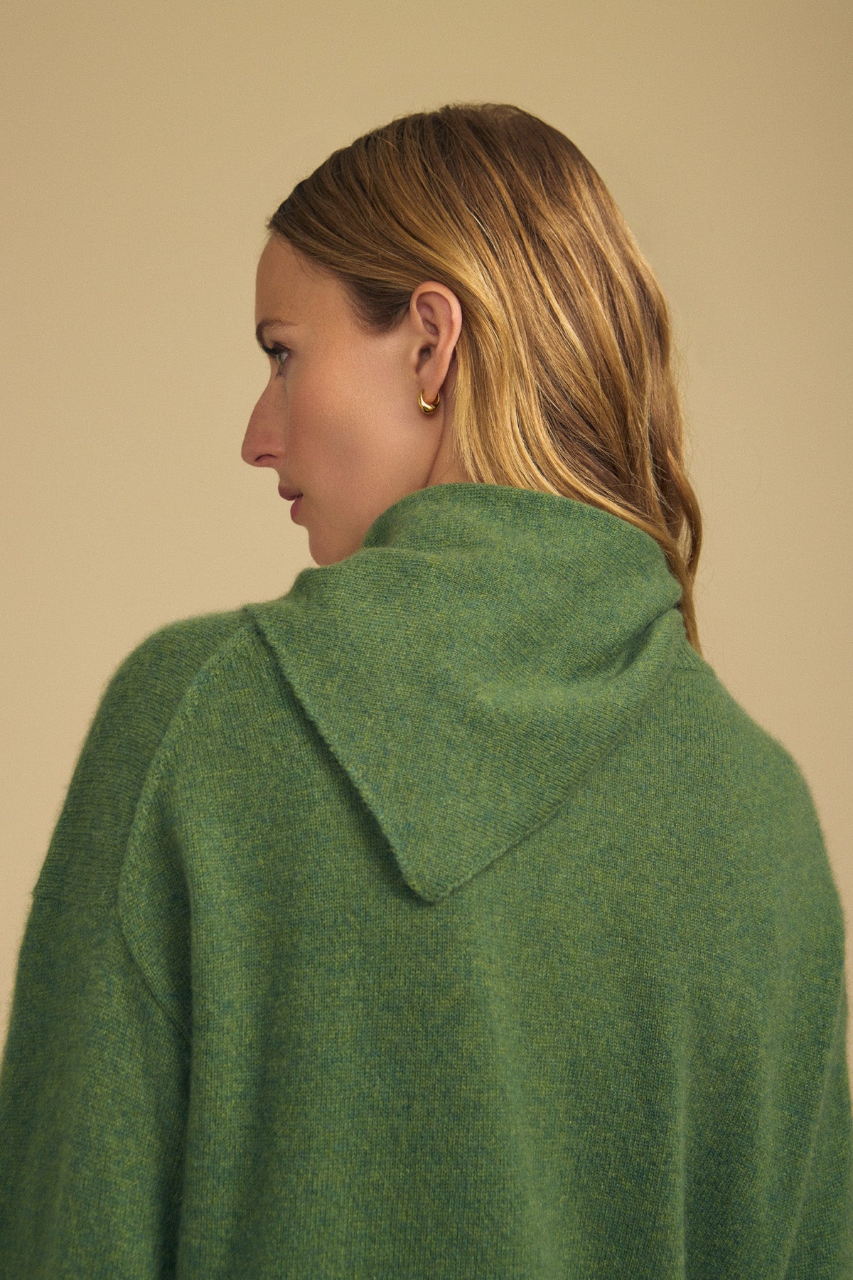A woman with light brown hair, wearing gold hoop earrings and a green hooded sweater, is shown from the back against a beige background. She accessorizes her look with the Velvet by Graham & Spencer HELEN CASHMERE SCARF, draped elegantly around her shoulders.-41960847343809