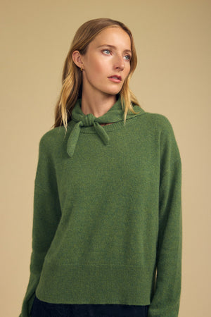 A woman with long, blonde hair wears a green sweater featuring the HELEN CASHMERE SCARF by Velvet by Graham & Spencer tied at the collar, standing against a plain beige background and gazing slightly to the side.