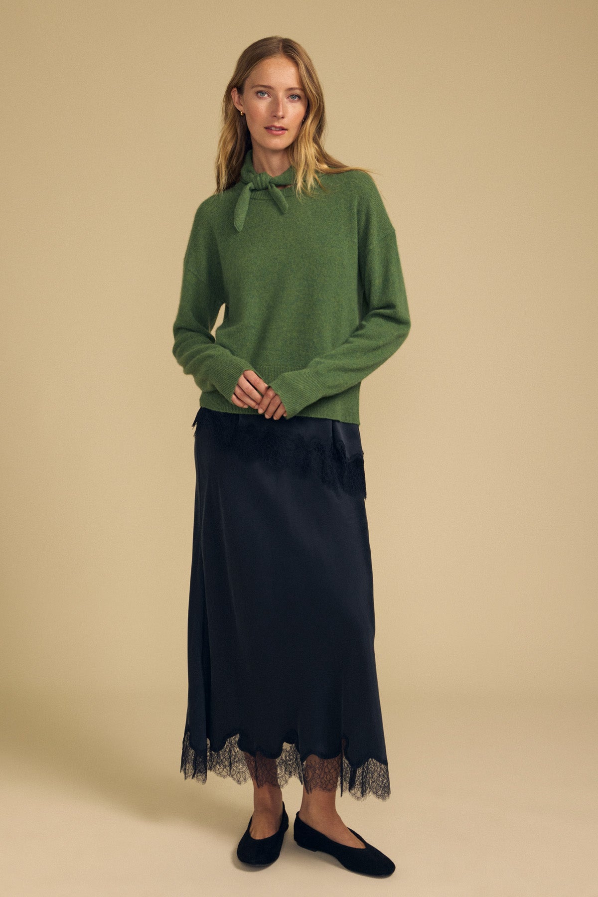 A woman stands against a plain beige background, elegantly layering her look with the HELEN CASHMERE SCARF by Velvet by Graham & Spencer draped over a green sweater featuring a bow at the neck. She pairs this ensemble with a long black skirt trimmed in lace and classic black flats.-41960847540417
