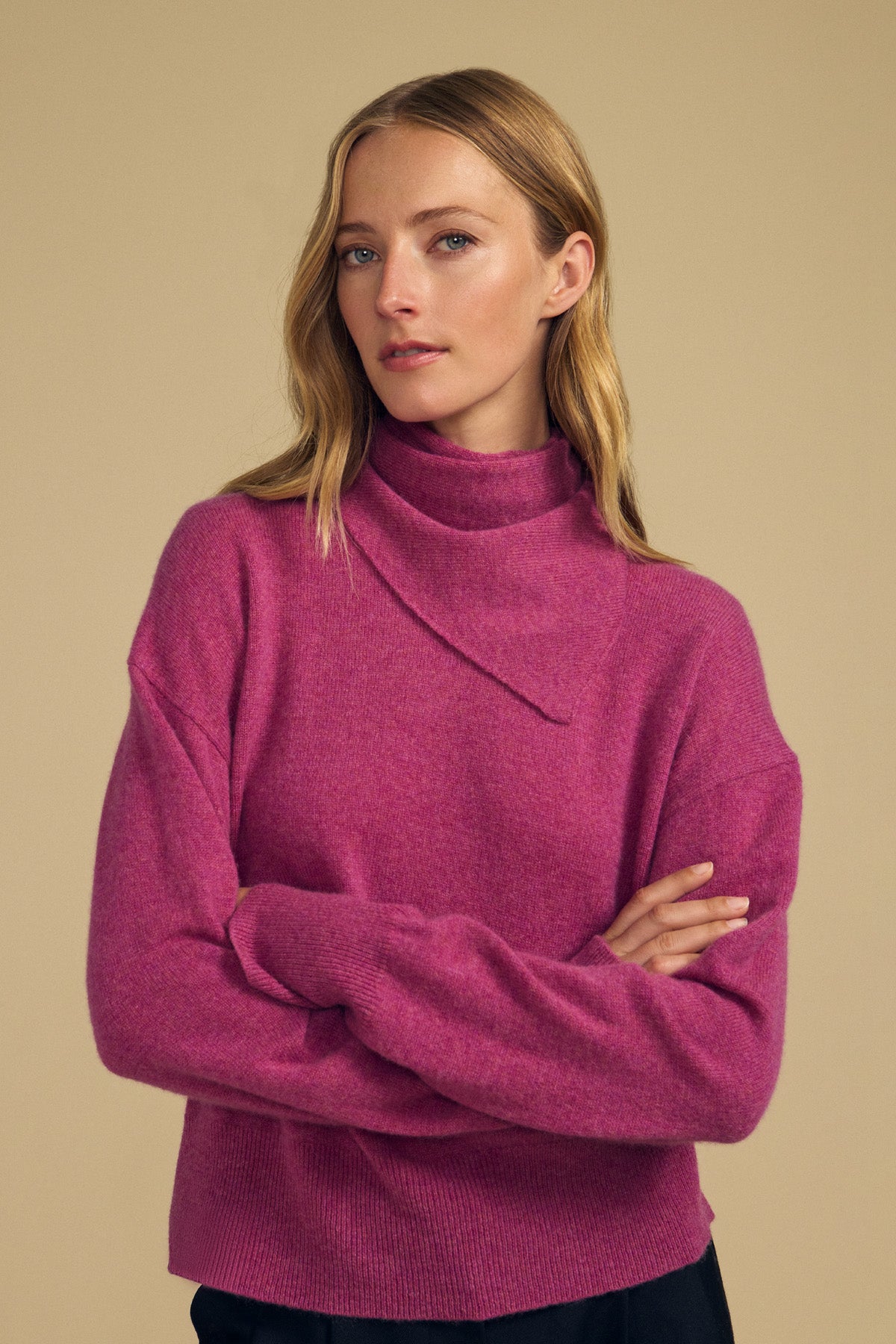 A woman with long blonde hair stands against a plain beige background with arms crossed, wearing a magenta turtleneck sweater and the HELEN CASHMERE SCARF by Velvet by Graham & Spencer.