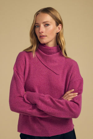 A woman with long blonde hair stands against a plain beige background with arms crossed, wearing a magenta turtleneck sweater and the HELEN CASHMERE SCARF by Velvet by Graham & Spencer.