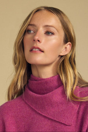 A woman with long blonde hair, dressed in a purple sweater and layering with the HELEN CASHMERE SCARF by Velvet by Graham & Spencer, stands against a plain beige background, gazing slightly to the side.