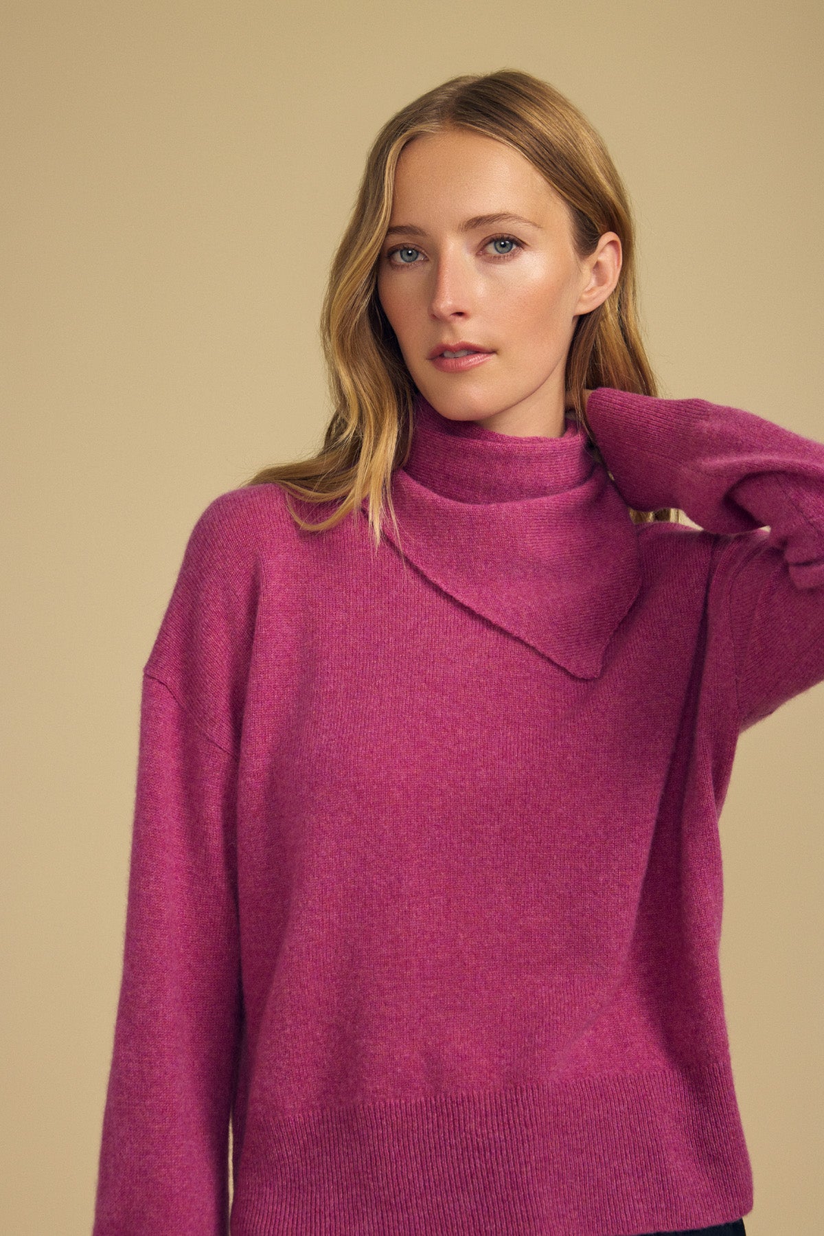 A woman with long blonde hair wears a magenta turtleneck sweater, effortlessly elevating her look by layering the Velvet by Graham & Spencer HELEN CASHMERE SCARF—a soft, triangular accessory—while standing against a plain beige background with one arm raised to touch her neck.