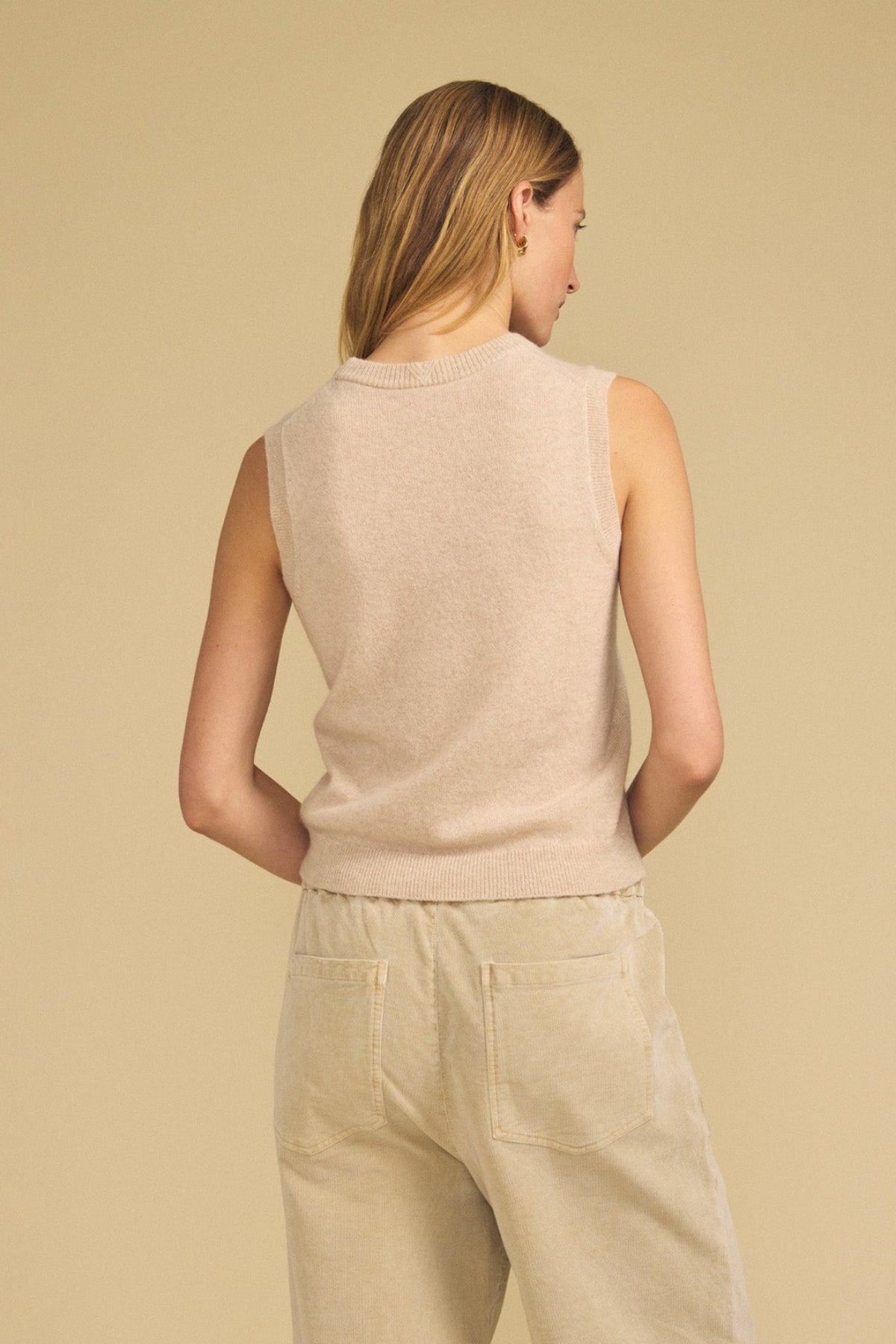 A person stands with their back to the camera, wearing the IVANA CASHMERE SWEATER VEST by Velvet by Graham & Spencer and light beige pants, set against a plain beige background.
