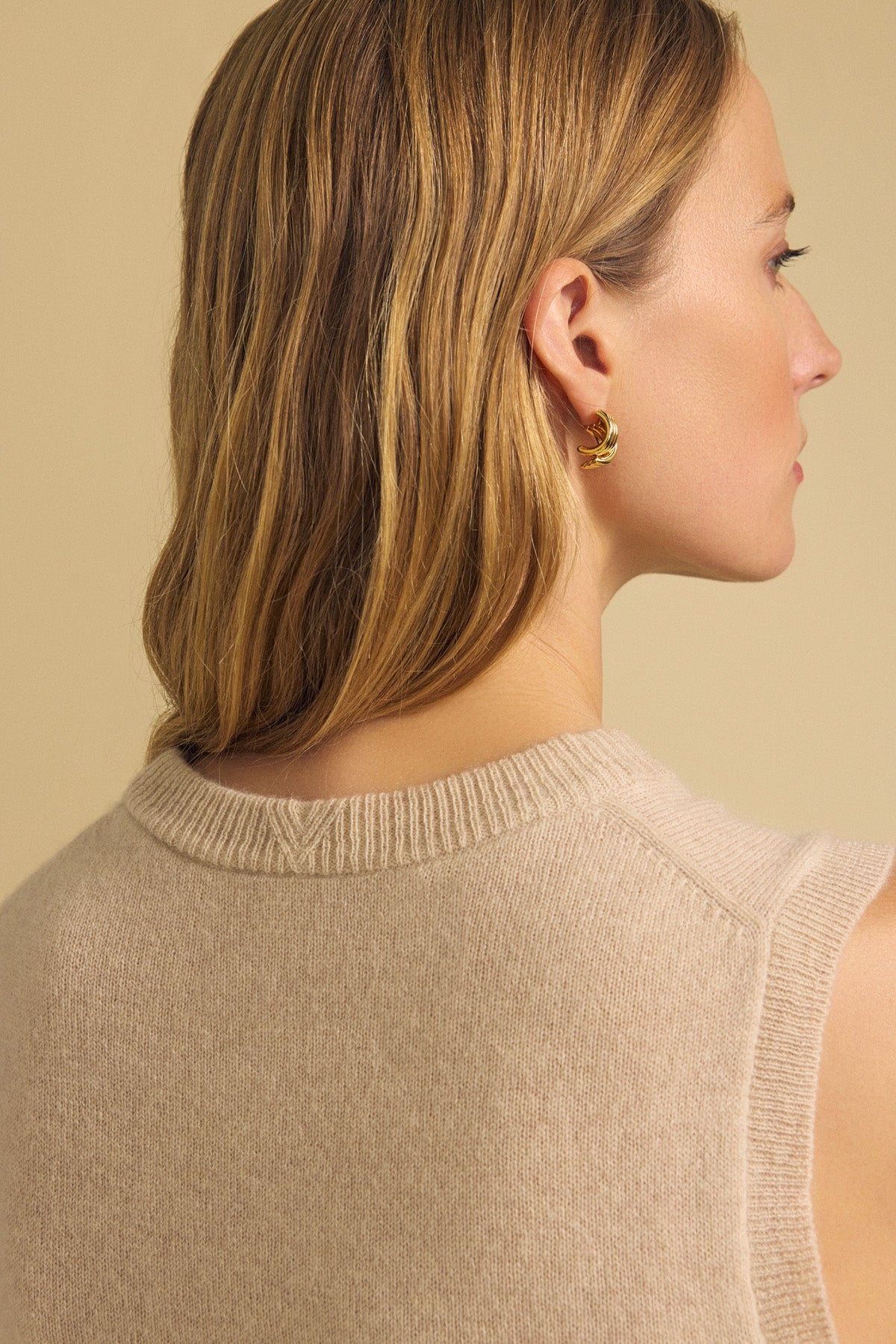 A woman with straight, light brown hair is shown from behind against a plain light background, wearing the IVANA CASHMERE SWEATER VEST by Velvet by Graham & Spencer and a gold hoop earring.