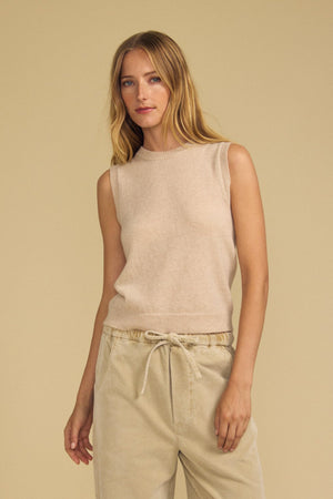 A woman with long blonde hair and a neutral expression stands against a beige background, wearing the IVANA CASHMERE SWEATER VEST by Velvet by Graham & Spencer, styled with beige drawstring pants.