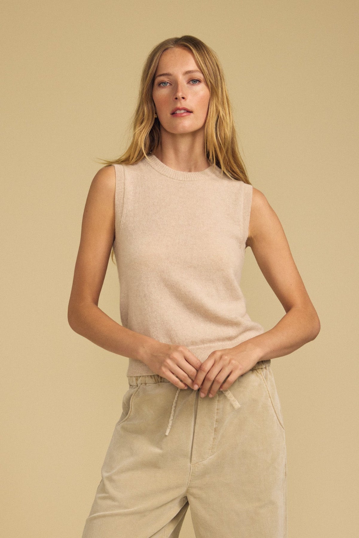 A person with long blonde hair wears the IVANA CASHMERE SWEATER VEST by Velvet by Graham & Spencer and light-colored drawstring pants, standing against a plain beige background.