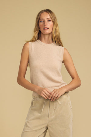 A person with long blonde hair wears the IVANA CASHMERE SWEATER VEST by Velvet by Graham & Spencer and light-colored drawstring pants, standing against a plain beige background.